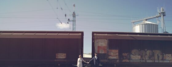 Yvonne und Klaus stehen Hand in Hand zwischen zwei Güterwaggons auf einem Industriegleis bei ihrem Hochzeitstrailer am Arche Moorhof.
