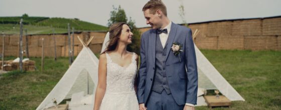 Jacqueline und Dominik stehen Hand in Hand vor den Tipis beim Holler Weingut Scheune, im Hintergrund die Weingärten – ein fröhlicher Moment aus ihrem Hochzeitsfilm.