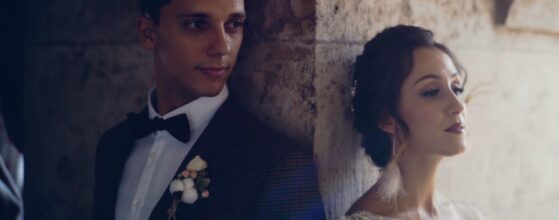 Nadya and Tomi leaning against the stone wall at Event’s Hill in Budapest, half in shadow and half in sunlight – a cinematic portrait from their Russian-Hungarian wedding film.