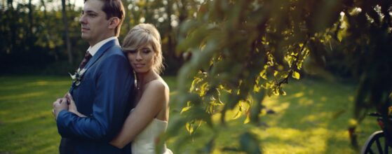Elisabeth umarmt Ingo von hinten im Garten der Schmiede am Ravelsbach, die Abendsonne scheint durch die Bäume – ein warmer Moment aus ihrem Hochzeitsfilm.