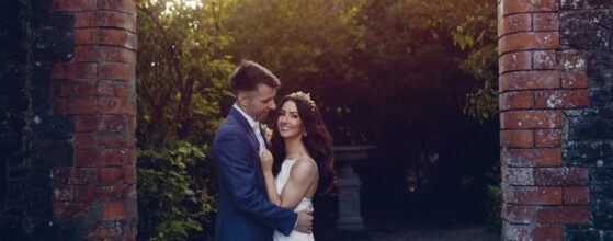 Caoimhe and Pauric standing close together between the brick arches at Larchfield Estate in Northern Ireland, with golden evening light around them – a romantic moment from their wedding film.