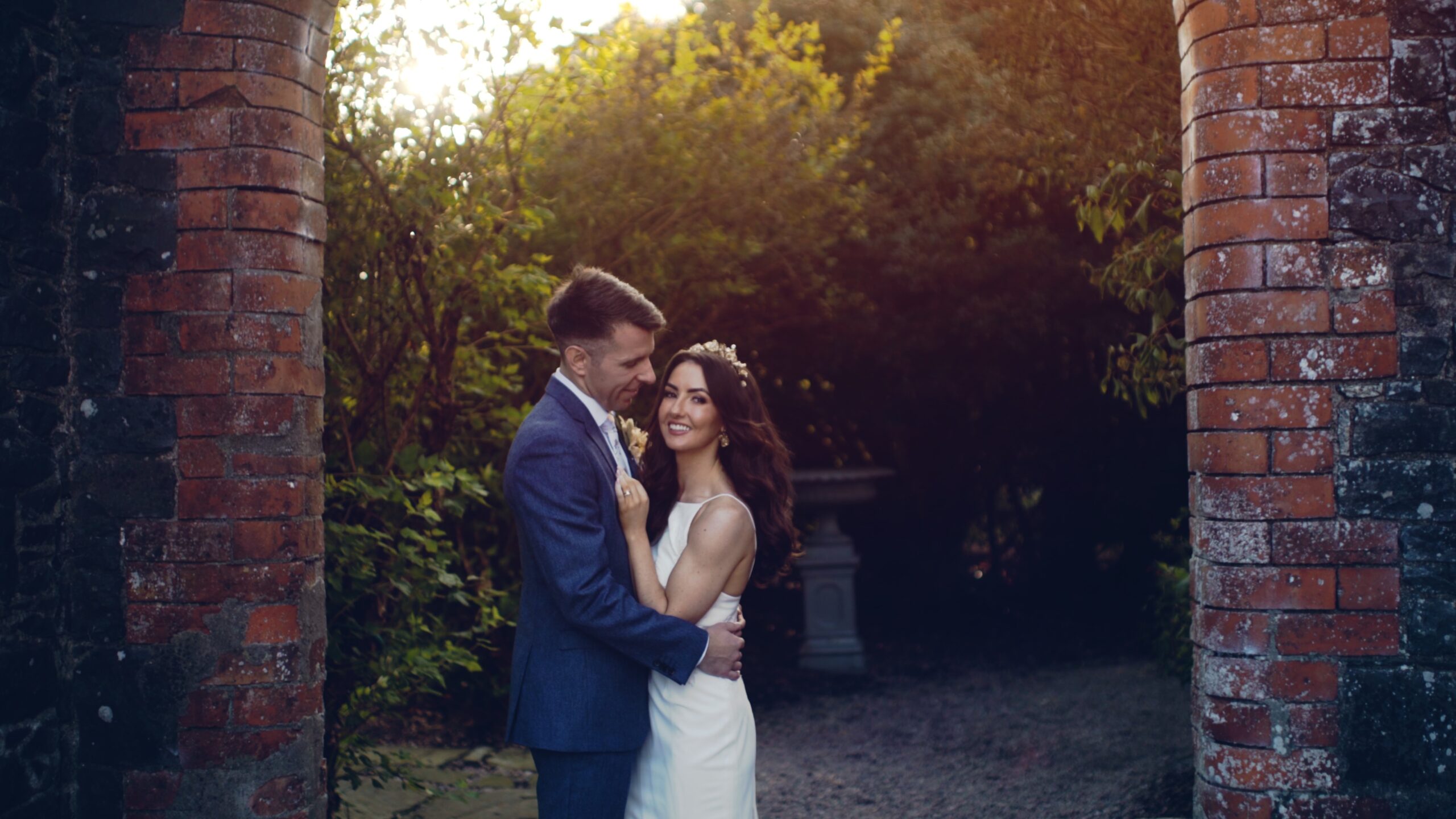 Caoimhe and Pauric standing close together between the brick arches at Larchfield Estate in Northern Ireland, with golden evening light around them – a romantic moment from their wedding film.