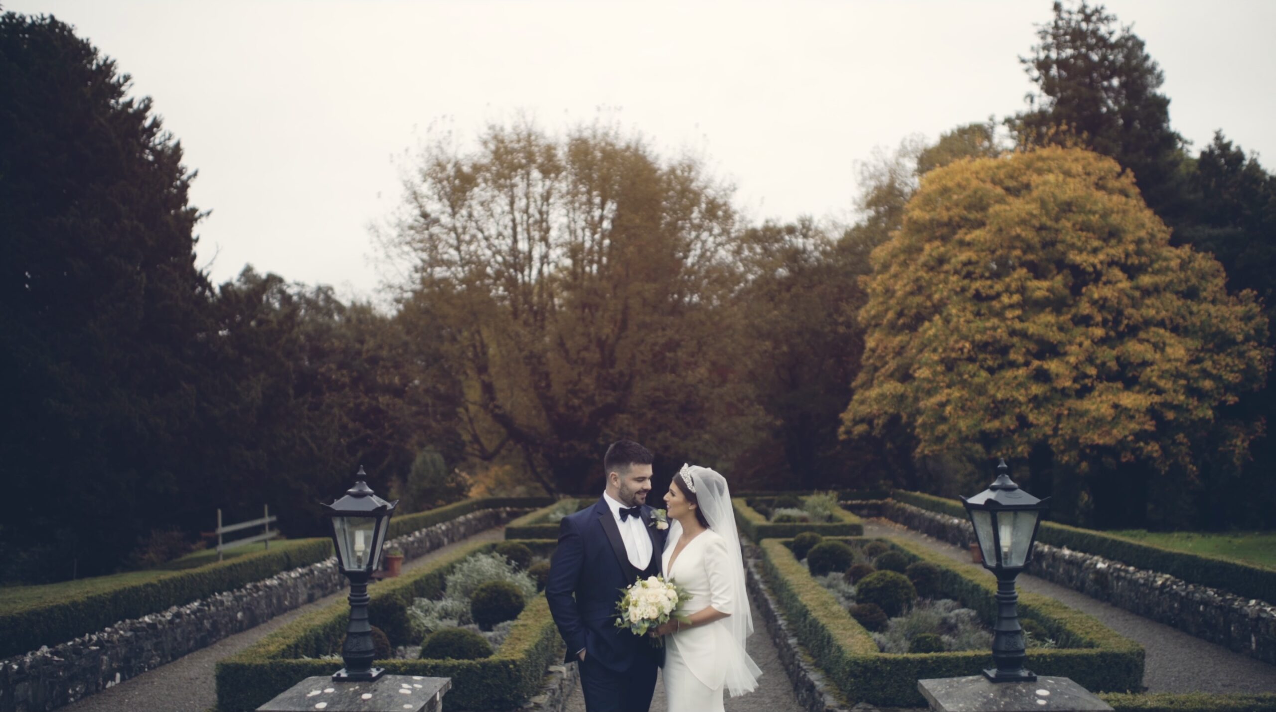 Clodagh and Peter standing together in the formal garden of Virginia Park Lodge in Ireland, surrounded by autumn colours – an intimate moment from their wedding film.