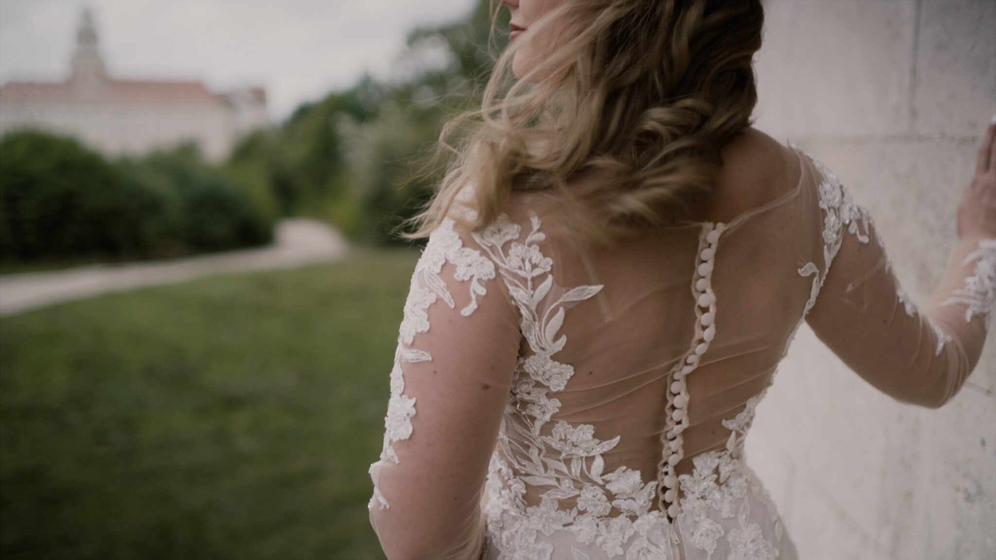 Szandra csipkés hátú menyasszonyi ruhában áll a pannonhalmi dombok között, finoman megérinti a falat – egy elegáns részlet az esküvői filmből. Pannonhalma esküvői film CulinArt
