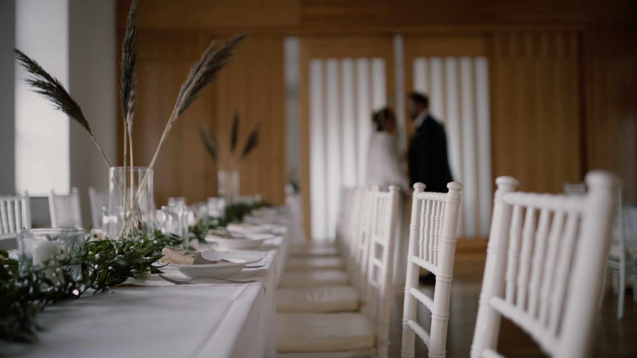 Hosszú, fehérre terített esküvői asztal Klebelsberg Kastélyban Budapesten, a háttérben Kata és Bali ölelkeznek – egy letisztult, elegáns részlet az esküvői filmből.