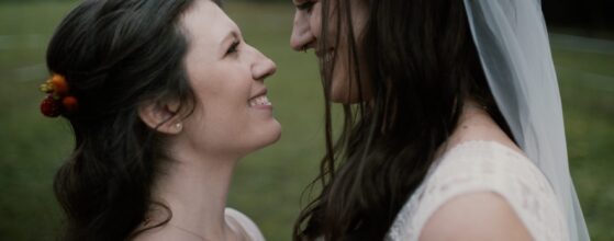 Sanny und Flo stehen sich lächelnd gegenüber auf der Wiese beim Lässerhof in Stattegg, zwei Bräute in Spitze – ein inniger Moment aus ihrem LGBTQ+ Hochzeitsfilm.