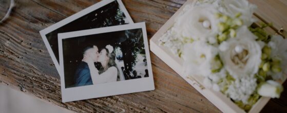 Zwei Polaroid-Fotos von Diana und Alex liegen auf einem Holztisch neben einer Blumenbox – ein verspieltes Detail ihres Hochzeitsfilms im Weingut Holler in Spielfeld.