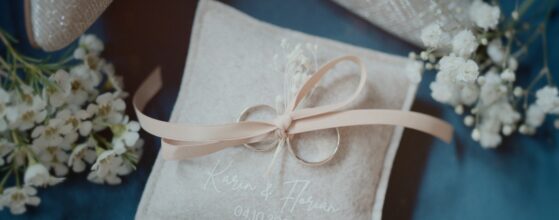 Eheringe von Karin und Florian auf einem personalisierten Ringkissen mit Datum, umgeben von zarten weißen Blüten – Detail ihres Hochzeits‑Teasers im Landgasthaus zur Krone in Gaaden.