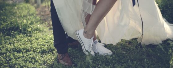 raut Anisja zeigt ihre weißen Converse-Sneaker unter dem Hochzeitskleid neben den eleganten Schuhen von Florian auf der Wiese in Spielfeld, Modern Casual Wedding Videographer Spielfeld Weingegend.