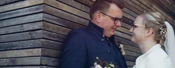 Barbara und Michael lehnen an der Holzfassade beim Obstbau HAAS in Gnas, sie sehen sich an und lächeln – ein natürlicher Moment aus ihrem Steiermark Hochzeitsfilm.