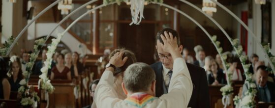 Pfarrer legt Corina und Martin während der kirchlichen Trauung die Hände auf den Kopf, im Hintergrund die Gäste – emotionaler Moment ihres Hochzeitsfilms im Winzerhotel Kolleritsch.