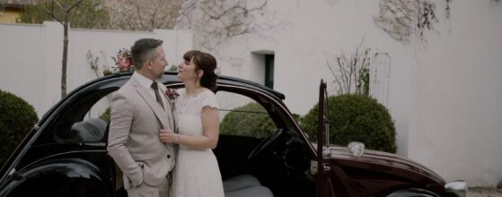 Dani und Alex stehen vor ihrem Oldtimer vor der Schmiede am Ravelsbach, sehen sich an und lächeln – ein entspannter Moment aus ihrem Hochzeitsfilm.