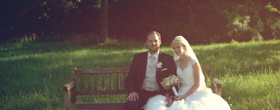 Daniela und Georg sitzen nebeneinander auf einer Holzbank im Park von Schloss Eckartsau, umgeben von Abendsonne und grüner Wiese Schloss Eckartsau Hochzeit