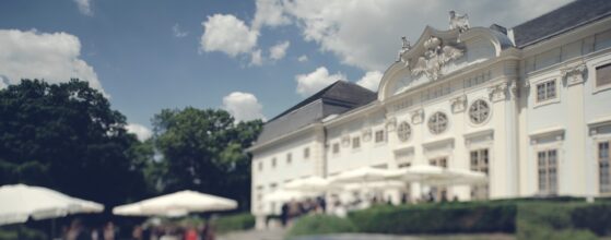 Luxury Castle Wedding Videographer Baroque Halbturn Hanna und Rainer stehen in der Schlosspark-Gartenanlage des Schloss Halbturn mit weißen Hochzeitszelte im Hintergrund und der eleganten Barockfassade des Schlosses