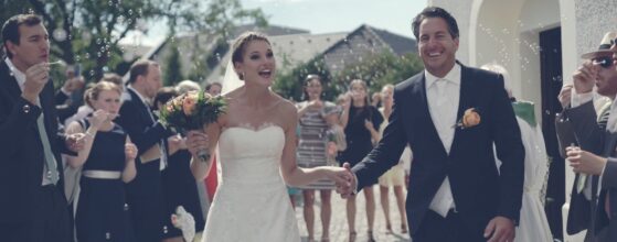 Jacqueline und Michael führen fröhlich ihren Hochzeitszug durch den sonnigen Hof der Jacky's Mühle in Gols – umgeben von lachenden Freunden und Familie im Burgenland