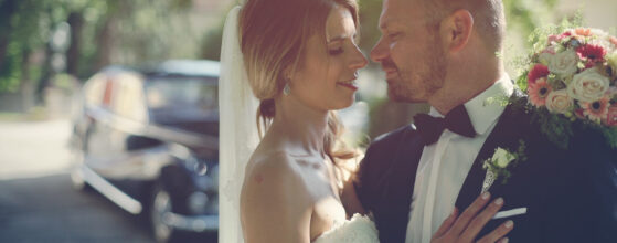 Jasmin und Florian stehen sich nah gegenüber, sie in Brautkleid mit Schleier und er im dunklen Anzug mit Boutonniere, hinter ihnen ein Auto. DER jung WIRT Hochzeit.