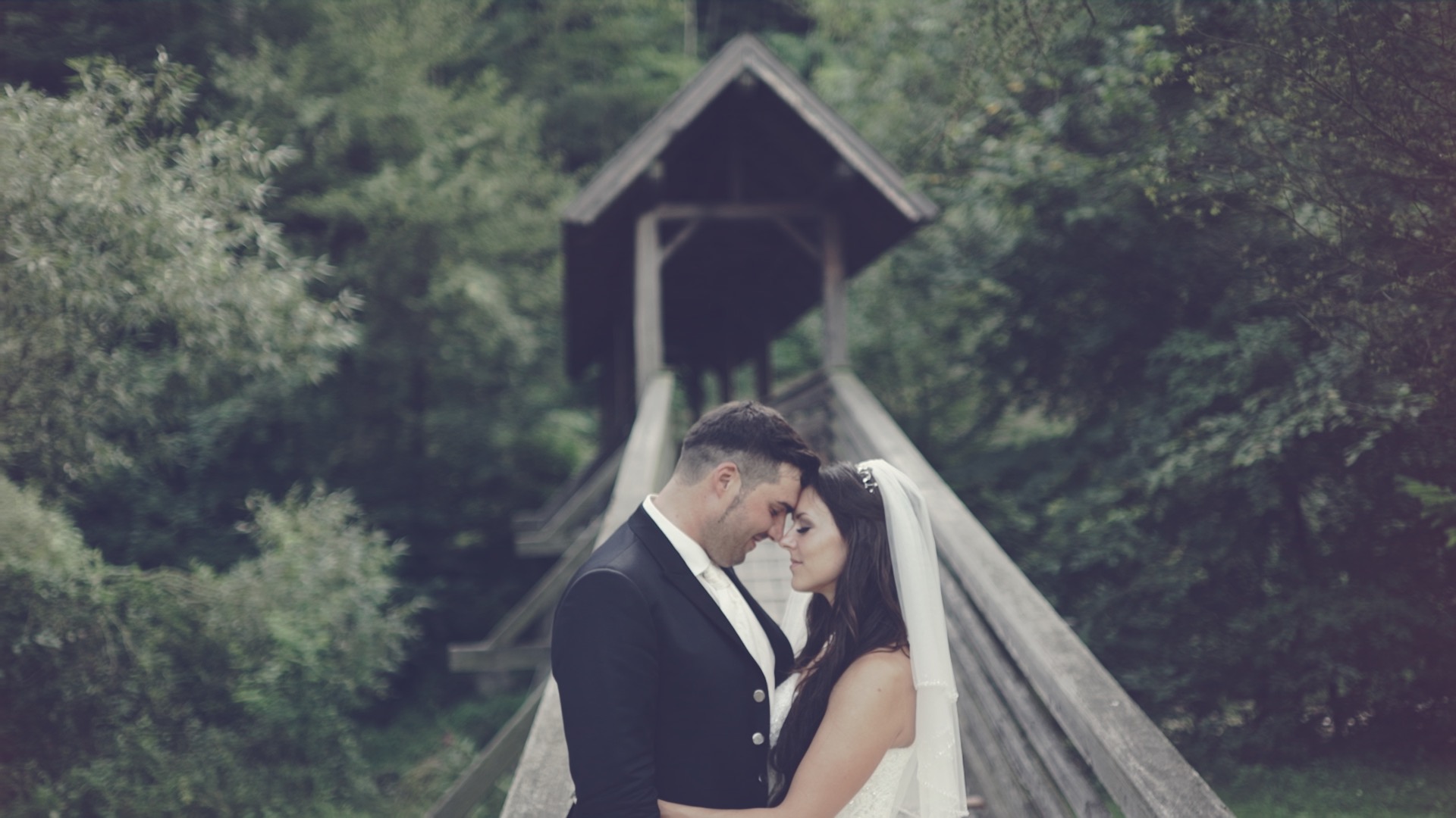 Jeannine und Manuel stehen eng umschlungen vor der überdachten Holzbrücke in der Natur der Krainerhütte bei Baden bei Wien