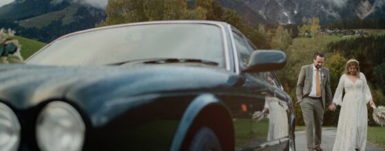 Jessica und Kevin gehen Hand in Hand neben ihrem Oldtimer durch die Berglandschaft von Leogang, im Hintergrund die Wolken über den Salzburger Alpen – Szene aus ihrem Hochzeitsfilm beim Der Kirchenwirt. Salzburg hochzeit videograf