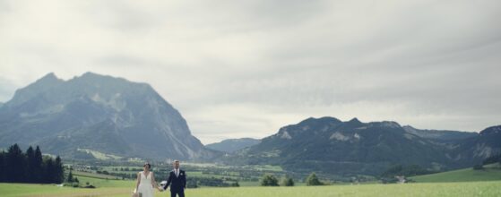 Braut Joanna und Bräutigam Andreas stehen hand in hand auf einer grünen Wiese vor dem Schloss Pichlarn mit dramatischen Bergen im Hintergrund – internationales Hochzeitsfoto.