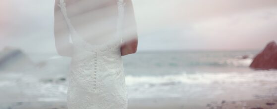 Bride Lani in flowing white lace wedding dress stands on the dramatic beach at Fort Polhawn, Cornwall, with crashing waves and red cliffs in the background during her wedding film