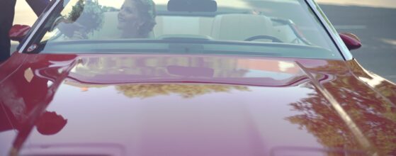 Maria sitzt im roten Cabrio vor Jacky’s Mühle in Gols, während Paul ihr den Brautstrauß durch die Frontscheibe reicht Jackys Mühle Hochzeit.