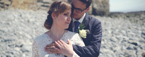 Marta and Mat stand close together on the rocky beach at Nash Point Lighthouse in Wales, Marta in white lace wedding dress and Mat in dark suit with white boutonniere
