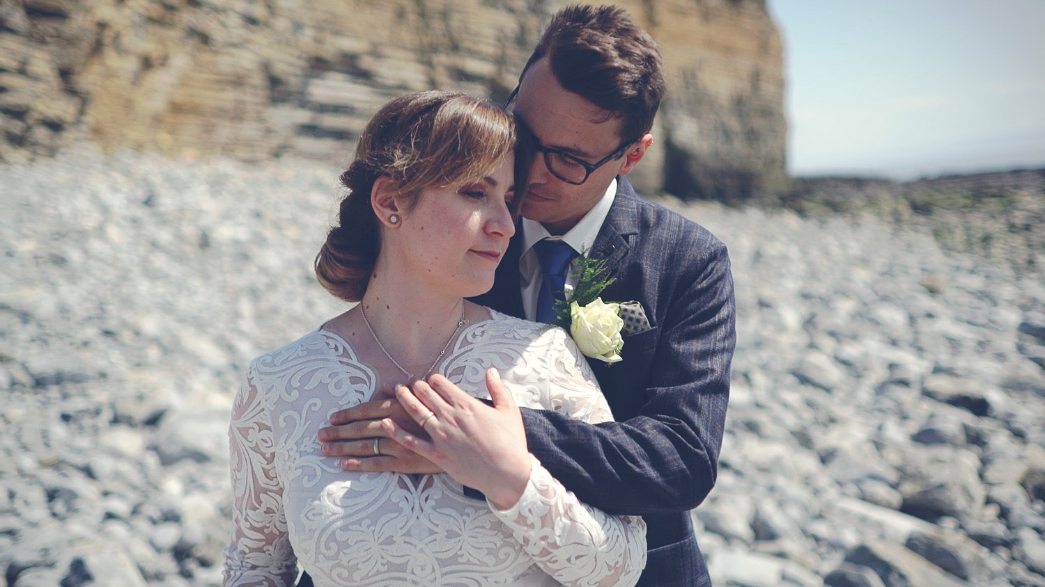 Marta and Mat standing together on the rocky beach near Nash Point Lighthouse in Wales, a relaxed seaside wedding portrait from their Wales wedding film.