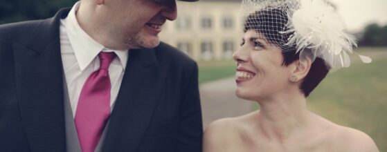 Monika in weißem Brautkleid mit Fascinator und Peter im dunklen Anzug mit rosa Krawatte lachen zusammen vor dem Schloss Laxenburg – romantisches Hochzeitsfoto