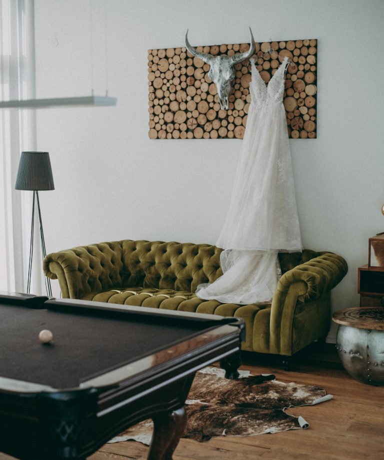 Brautkleid hängt im modernen Lounge-Bereich der Holler Scheune auf einem grünen Samtsofa – entspanntes Getting Ready in der Hochzeitslocation Weingut Holler in der Südsteiermark. Holler Weingut Hochzeitsfilm Spielfeld