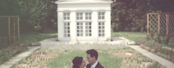 Ancsi és Tomi egymás mellett állnak a Rókusfalvi Birtok fehér neoklasszikus pavilonja előtt, zöld ösvénnyel körülvéve az esküvői képen.